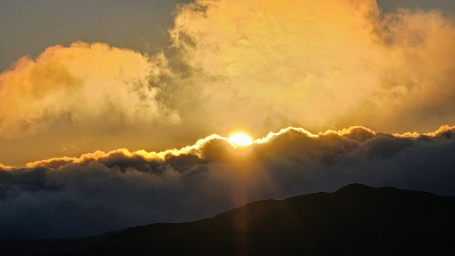 A vibrant sunset captured with the sun bursting through a horizontal band of golden-orange clouds, casting rays of light downwards. Below the clouds, the dark silhouette of a mountain range stretches across the bottom of the frame. The upper portion of the sky is filled with more warm-toned clouds, hinting at the expansive and beautiful skyscape.
