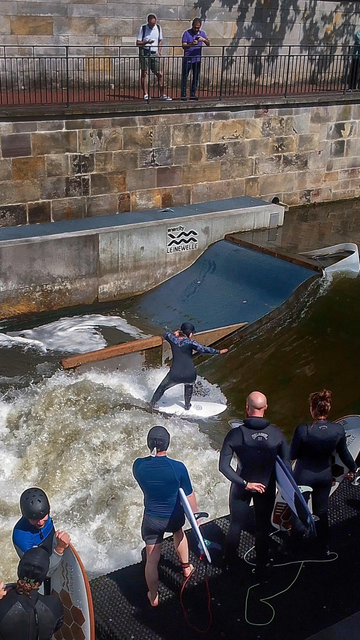 Blick auf die Leine, Innenstadt Hannover. Es ist sonnig. Eine Frau auf einem Surfbrett surft auf der sogenannten Leinewelle, eine künstlich angelegte Strömung zum Surfen. Am Ufer vier weitere Personen mit Surfbrettern, auch in Neopren-Gummianzügen und teilweise mit Helmen, sie warten am Einstieg. Hintergrund: das gemauerte Ufer. Oben stehen zwei Zuschauer. Das Wasser ist grün und schäumt weiß.