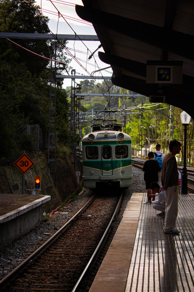 The "frog" train entering Les Planes station on platform 1, about to head towards Terrassa. There's a speed limit sign and a red traffic light on the left.