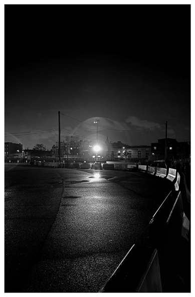 Monochrome photo of a wide stretch of empty pavement at the Brooklyn Cruise Terminal, at night. A line of low traffic barriers in the foreground stretches away towards right frame, then swerves to the left in the middle distance and disappears at left frame. In the center of the photo, a single streetlight gives off a halo of concentric circles and is reflected in a small puddle in front of the barriers. Beyond the streetlight are electric poles and a row of low-rise buildings with lit windows. Dim clouds float in a gray sky that fades to black near the top of the image.