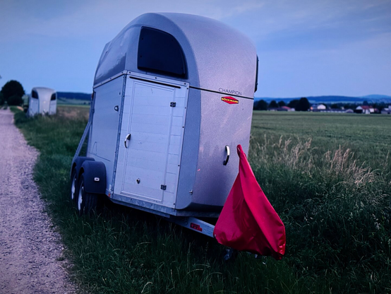 A silver horse trailer is parked on a gravel road next to green fields. A red flag is attached to the rear of the trailer. The sky is slightly overcast, suggesting twilight or dusk. In the background, a second horse trailer is visible along