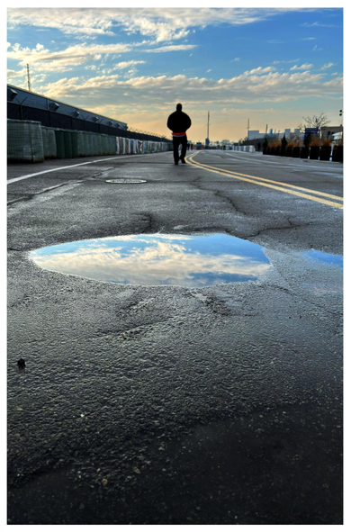 A wide stretch of pavement flanked by low buildings, at the Brooklyn Cruise Terminal in the late afternoon. A puddle in the foreground reflects the clouds and vibrant blue sky above. Pavement markings — a white line at left frame, a double yellow line at right frame — recede in a curved path and converge in the distance. A solitary figure, wearing a dark jacket with a red shirt peeking out from the bottom, walks away from camera in the center of the image. 