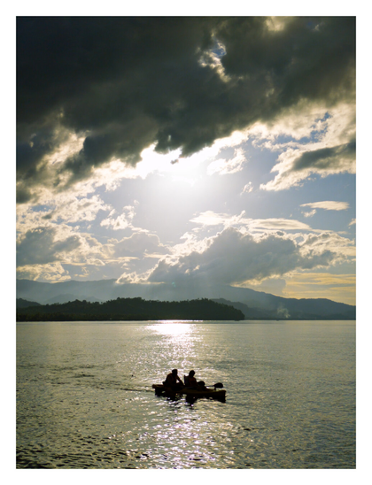 A dramatic, backlit shot shows two people in a small boat on a shimmering body of water. Above them, the sun breaks through dark, heavy clouds, with hazy mountains visible in the distance.