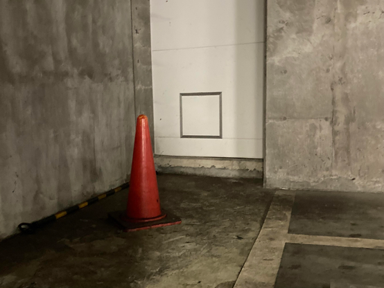 A bright orange traffic cone stands in a dimly lit concrete area, next to a white wall with a square inset. The floor shows signs of wear, and a yellow and black striped barrier is visible along the lower edge.