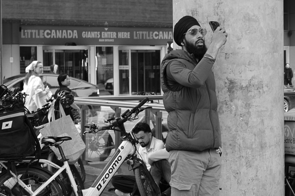 The right half of the frame is filled by a man wearing a turban, sunglasses and a puffer vest holding up his phone up high to record something he’s watching. Behind him is the concrete texture of a support column. Behind him, in the lower left are the bicycles of food couriers, with one rider sitting on the ground looking at his own phone. In the far background are pedestrians, and the signage on the building across the street.