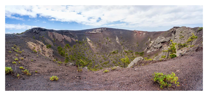 The picture shows the San Antonio volcano on the island of La Palma. It is a large, crater-shaped volcano with steep, sloping walls consisting of dark volcanic soil and rock. The crater is visible from above and has a broad, oval shape. Scattered green plants and small trees grow on the slopes, standing out against the dark soil. The sky above is partly cloudy, with patches of blue shimmering through the white clouds.
