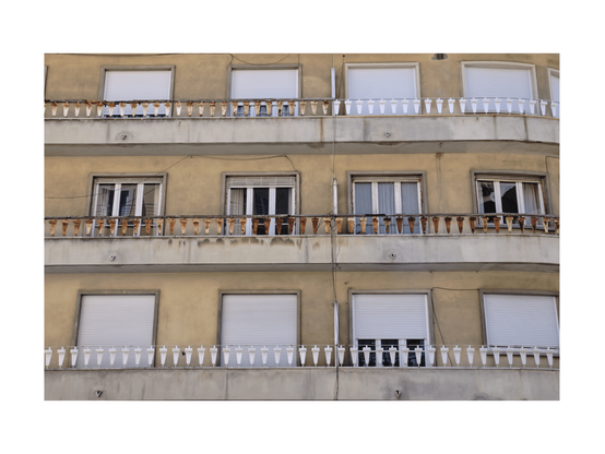 close up of a residential building with yellowish facade in a lisbon neighborhood. most shutters are down and parts of balcony railings are rusty and white.
