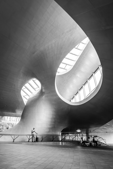 Black and white photo of the futuristic interior of Arnhem Central Station in the Netherlands, featuring sweeping, organic architectural curves and large oval skylights that let in natural light. A lone traveler with a suitcase stands near a railing on the left, while three people sit on steps to the right beneath the structure. The flowing ceiling design creates a sense of movement and space, emphasizing modern architectural aesthetics.