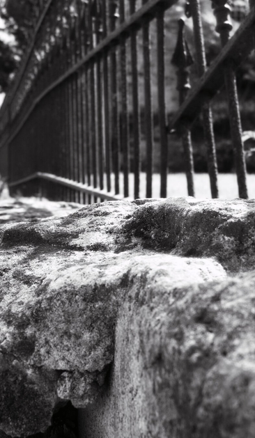 A black and white photo showing a low angle view of a stone wall with a black metal fence.
