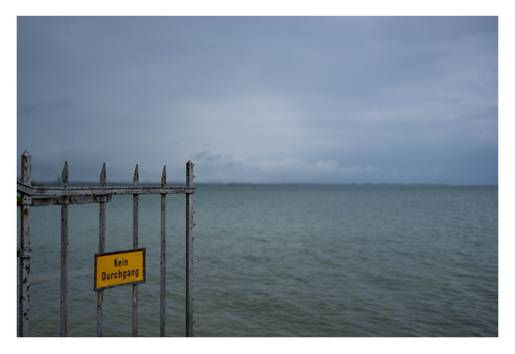 Foto im Querformat. Blick auf einen großen See. Der Horizont ist genau mittig im Bild, der Himmel bewölkt. Links ragt Teil eines alten Metalltores ins Bild. Der obere Teil ist parallel zum Horizont, die Spitzen ragen darüber. Auf dem Tor ist ein gelbes Schild „Kein Durchgang“.