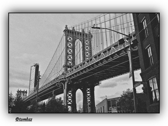 A black-and-white photograph of the Manhattan Bridge in New York City. The view from the street between buildings draws the eye toward the majestic steel structure, whose arches and cables form a distinctive silhouette against the sky.
