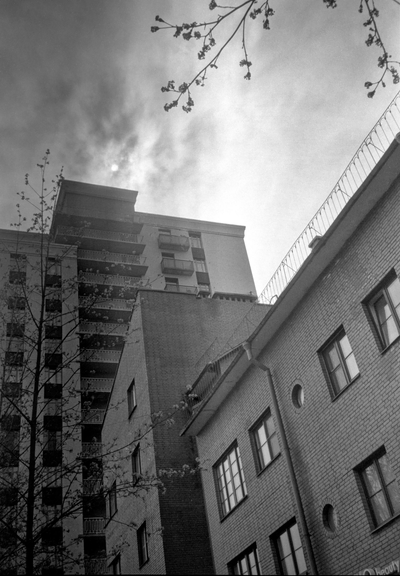 Monochrome photo shows two high-rise buildings, above which the disk of the sun behind the clouds is visible.