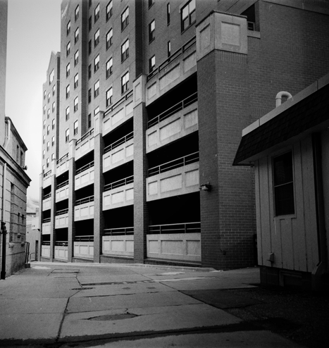 Photograph, black and white. View of an urbanized back alley, with a four-story brick and concrete parking structure centered. 