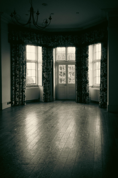 Inside a large room. A wooden floor, reflecting the light, looking towards a large bay window and door.