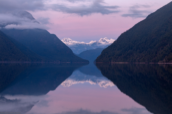 A reflection of mountains in a lake after sunset - with a purple sky.  Mountains on the left and right are close by, covered in trees, and relatively low elevation.  In the middle of the frame are more distant, rocky, mountains with some fresh snow on them.