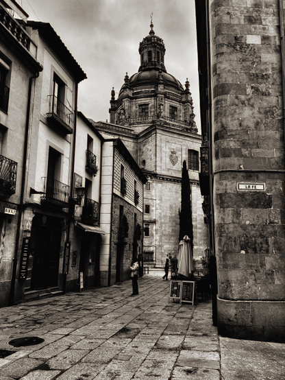 Una calle estrecha y empedrada en blanco y negro muestra edificios antiguos a cada lado, con balcones de hierro forjado. Al fondo, se alza imponente una gran cúpula de una iglesia o catedral de estilo barroco, mostrando su arquitectura detallada. Una persona se encuentra de pie en la calle, cerca de un café con un parasol. La escena evoca una atmósfera histórica y tranquila.