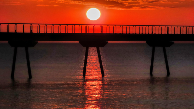 La imatge mostra una silueta d'un pont sobre el mar al capvespre. El sol gran i brillant es troba just darrere del pont, pintant el cel i l'aigua d'un color vermell ataronjat intens. La silueta del pont contrasta amb la lluminositat del cel, creant una imatge dramàtica i potent.