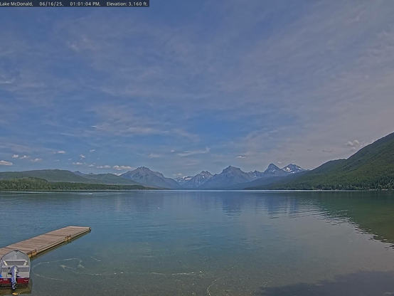 Calm water, an empty dock, and distant mountains in this view from Lake McDonald in Glacier National Park today. A single boat remains tied to the dock. A variety of cloud shapes appear in an otherwise blue sky.  The slopes surrounding the lake are green with vegetation. Snow dots the mountain peaks. 