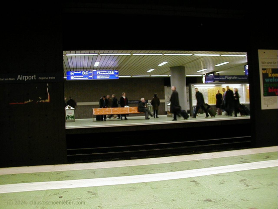 The subway station in Frankfurt(Main Airport (Germany). About a dozen persons in suits and black coats and with trolleys are waiting for or running hastily to their connections.

The picture is taken from the empty platform on the other side of the railtracks and it seems you are looking into a kind of theatre stage playing an absurd scene.

