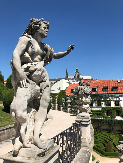 A statue of a nymph looks down into the sculpted gardens below. The sky is bright blue. In the background are white buildings with red rooftops 