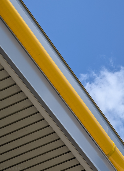 The composition creates strong diagonal lines and plays with contrasting textures and colors – the smooth, vibrant yellow against the cooler, more textured greys and whites, all set off by the clear blue sky. 