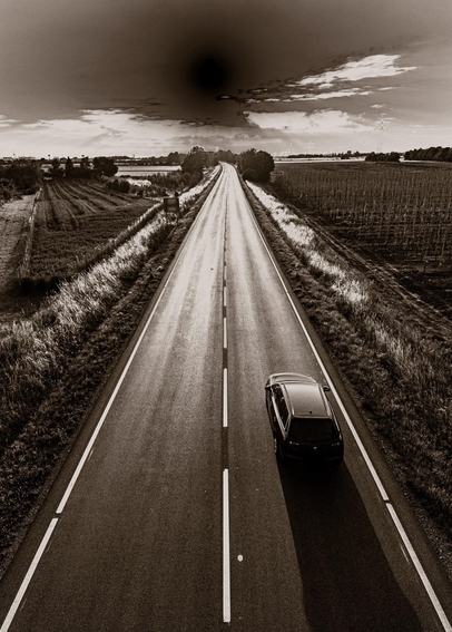 A sepia toned monochrome image of a road from a bridge over it. The road is straight, curving slightly left in the distance, under the late evening sun. The intensity scale has been inverted at the bright end, turning the sun in to a black hole. There is a car on the right hand side of the road in the foreground, heading away from the bridge and towards the sun. There are open fields on either side of the road.