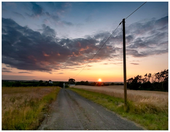 A gravel road leads through a field at sunset, with the sun dipping below the horizon. The sky is filled with colorful clouds, and a utility pole stands alongside the road. In the distance, a farmhouse is visible among trees.