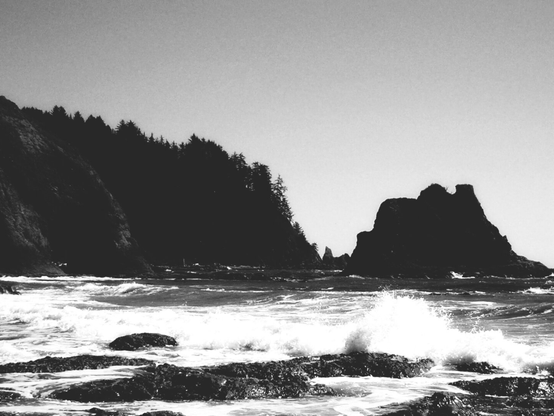 A black and white photo showing waves pounding a rocky beach and headland.