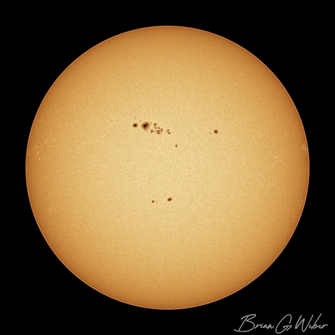 A full disk monochrome image of the sun colorized to orange with a thick cluster of sunspots on the upper middle part of the surface.