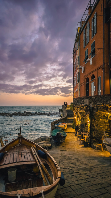 A romantic coastal scene at sunset, showing a charming, narrow stone walkway beside the sea. Several small wooden boats are moored along the edge, with one in the foreground featuring wooden oars and a bucket inside. The right side of the image is dominated by a tall, orange building with green shutters and laundry hanging from windows. Golden lights softly illuminate a cozy outdoor seating area with potted plants and wooden chairs, leading up stone steps to two silhouetted figures gazing out over the water. The sky is painted in soft purples and oranges as the sun sets behind dramatic clouds, casting a warm glow over the tranquil sea.