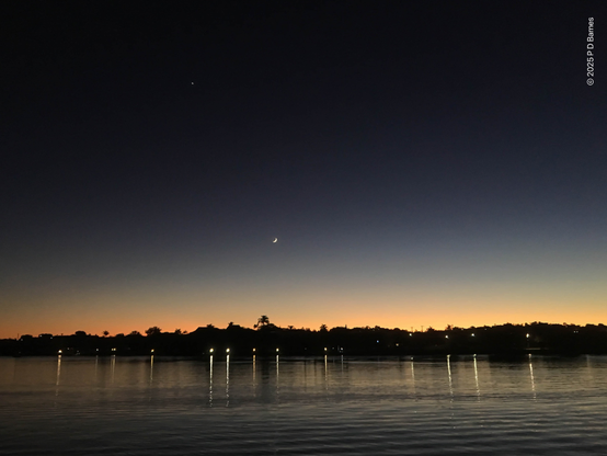 Looking across a calm Brisbane river at dawn. On the opposite bank the land is still dark, lights on the riverwalk reflecting in the river, trees silhouetted against the early orange glow just creeping over the horizon. Higher up a small crescent moon hangs in a slate-blue, almost black-ink sky.