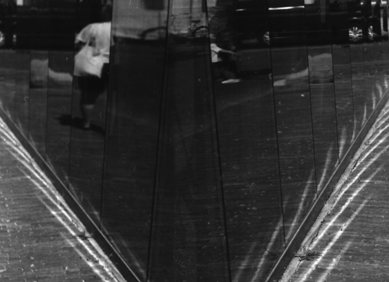 A corner of a glass building where the sun creates a pattern on the ground, and the glass reflects a busy street.