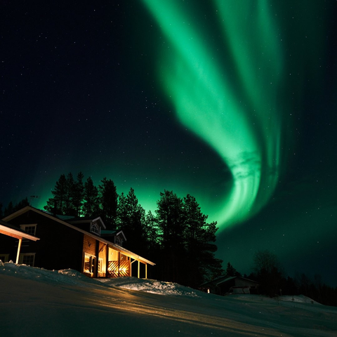 The aurora streaks across the night sky above a wooden cabin and a treeline behind the cabin in a snow covered landscape.