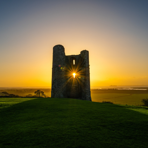 The ruins of one of the towers of Hadleigh Castle at sunrise. The sky is a gentle gradient of orange-yellow, turning more blue towards the top of the photograph. The sun shines through one of the windows in the wall of the ruined tower.