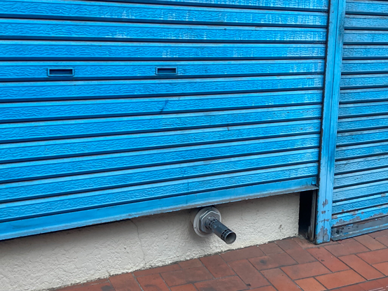 A blue metal shutter partially covers a wall, with a ventilation pipe protruding from the bottom. The wall itself is textured and painted white, with a tiled floor visible in front.