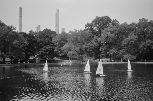 Four model Laser class sailboats ply the waters of the tree-lined Conservatory Water. Several of the City's supertall apartment buildings stand in the distance. Rentals are $15 for 30 minutes--for the boats, not the apartments.