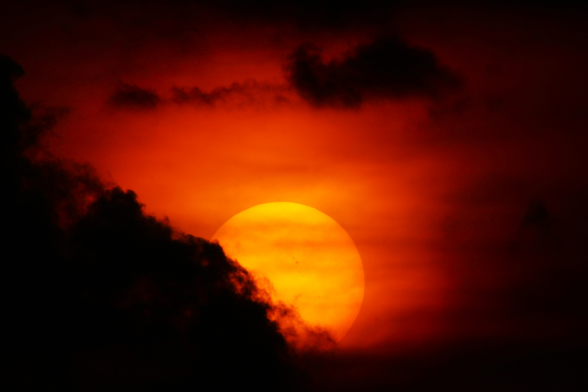 Sunset photo of a large yellow sun in orange clouds with a dark cloud from the lower left moving across it to cover its lower left. Sunspots are visible on the Sun.