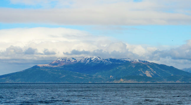 on the horizon, a massive mountain rises from the sea to snow caps, where it disappears into clouds under an ethereal blue, grey, white sky
