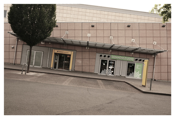 Straßenansicht eines modernen Gebäudes mit einer rosa-braunen Fassadenverkleidung und einem leicht geneigten Vordach. Links befindet sich ein Eingang zur „Postbank Finanzcenter“-Filiale mit gelbem Rahmen. Rechts daneben liegt ein „Sonnenstudio“ mit grüner Beschriftung und zwei gläsernen Türen. Zwischen den Eingängen steht ein Baum mit Fahrradständer. Der Himmel ist nicht sichtbar, der Fokus liegt auf der Fassade.