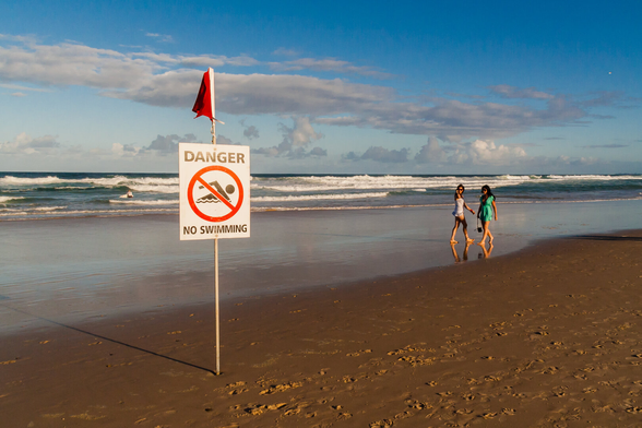 A beach scene with a danger, no swimming sign on the left and two young women in the distance on the right.