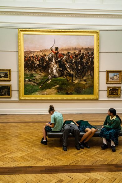 Four schoolchildren relaxing in an art gallery. The two in the middle, on their backs, apart to be looking at the painting on the wall behind them, upside down.