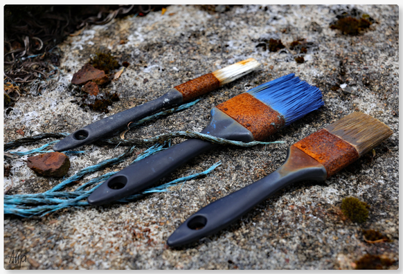 Drei Pinsel liegen sorgsam aufgereiht auf einem Felsbrocken: die Farben an den Pinselköpfen sind blau, weiß und braun und eingetrocknet. Die Verbindungen zu den dunkelblauen Stielen sind verrostet. Rostrot sind auch die Moose und Steinchen auf dem Stein.  

Three brushes lie carefully lined up on a boulder: the colours on the brush heads are blue, white and brown and dried out. The connections to the dark blue handles are rusty. The moss and pebbles on the stone are also a rusty red colour.