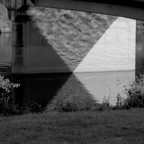 A black and white image of a concrete footbridge over a stream. The dusk light reflects on the water in such a way as to make a perfect triangle on the bridge column with the light, shadow, and reflection of the caustics on the water's surface