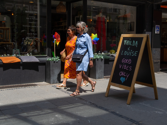 The image depicts two women walking on a sidewalk in front of a store. The woman on the left is wearing an orange dress with a pattern, white sandals, and a brown handbag. The woman on the right is dressed in a light blue dress, black sandals, and a black crossbody bag. A chalkboard sign in front of the store reads "THILDA x LOUISE GOOD VIBES ONLY" with a blue heart symbol.
