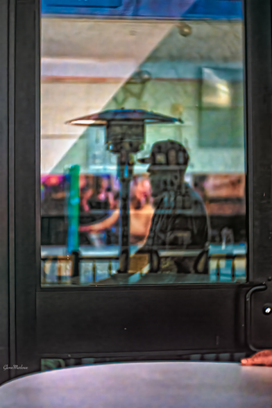 A blurred view through a door showcases a person beside a patio heater, with reflections of colorful lights and other patrons visible in the background of a social space.