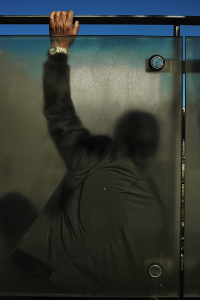 le dos d'un homme, le bras levé, la main tenant la barre d'acier de l'abribus, derrière une vitre sur laquelle il est appuyé