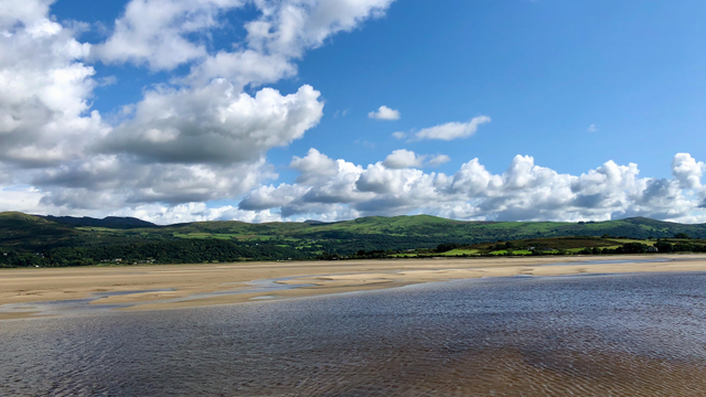 This picture showcases a serene coastal landscape with a wide stretch of sandy beach in the foreground, partially covered by shallow pools of water. Beyond the beach, there's a lush green expanse of rolling hills and farmland, dotted with patches of trees. The sky above is vast and bright blue, filled with fluffy white clouds casting gentle shadows on the land below. The scene exudes a peaceful, natural beauty, capturing the tranquility of a coastal countryside.