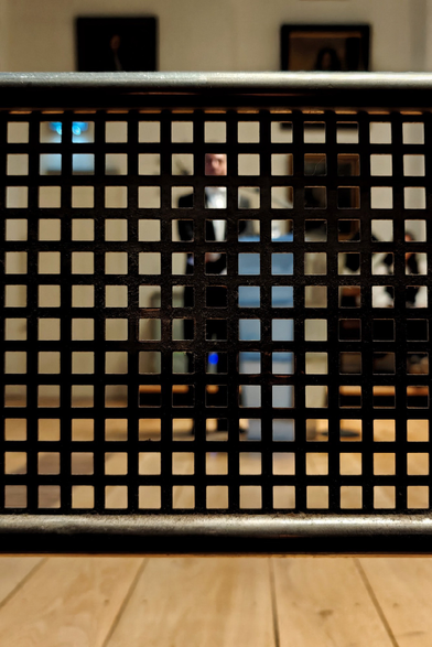 Looking through a black metal grid or grate with square openings one can see a room with wooden floor and cream-colored walls with framed pictures  in the background. A person in formal clothing is standing next to a small lectern, looking forward, their hands behind their back.