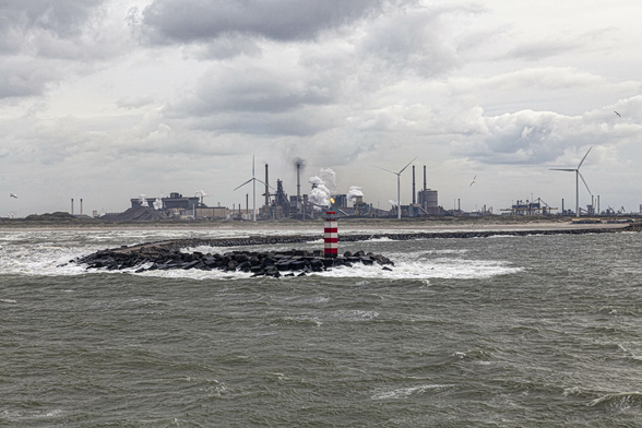 A gloomy, moody, deeply atmospheric view across a cold rolling sea of grey-green waves rippled heavily and topped with white flecks of foam. We're looking towards land which starts with a curving breakwater of black rocks and stones, topped with a narrow flat path that leads to the short, square, red-and-white-striped lighthouse on the end nearest us. Way back from this is the mainland and it's a flat one dominated by a huge industrial complex. Mounds of excavated material next to silos in front of squat, functional buildings, interspersed with towering chimney stacks. Cooling towers belch out thick white clouds of steam and a gas outlet pipe is lit with a steady flame. Evenly spaced, three wind turbines are just the tallest of the structures against the thick mass of grey cloud in the sky.