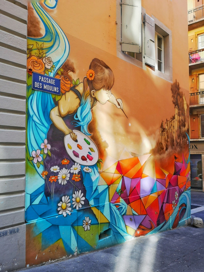 Das Bild zeigt eine Wandmalerei an einer Hauswand in einer engen Gasse. Sie ist farbenfroh und zeigt ein Mädchen mit teils langen, blauen Haaren. Es hält eine Palette mit Farben und  malt mit einem Pinsel. Es trägt ein dunklviolettes Kleid. Um es herum sind farbige Blumen und geometrische Figuren. Ganz rechts ist ein Schloss sichtbar. Das Bild wurde in der Passage des moulins gemalt, wie das angebrachte Steassenschild verrät.